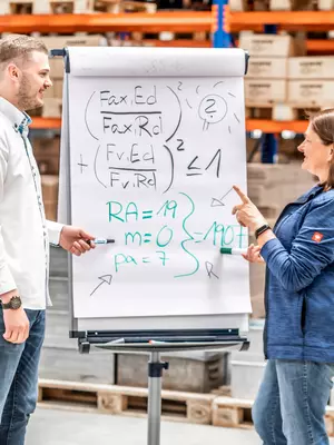 Zwei RAMPA-Mitarbeitende stehen nebeneinander in der Lagerhalle. Zwischen ihnen steht ein beschriebenes Flipchart, über dessen Inhalt sich die Mitarbeitenden austauschen. Das Bild soll die gemeinsame Ideenentwicklung symbolisieren und für RAMPAs Wert "Ein