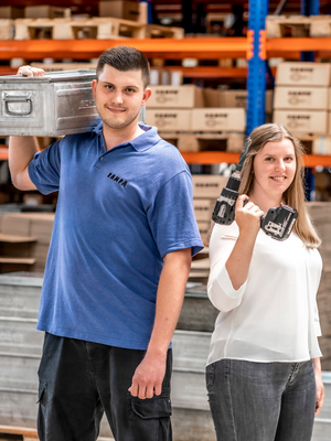 Two RAMPA employees stand next to each other in the warehouse. One is carrying a crate on his left shoulder, the other is holding a drilling machine in her hand. The picture is supposed to stand for RAMPA's value "tackling".