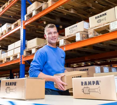 A RAMPA employee from the warehouse holds a RAMPA carton. In front of him are more cartons, behind him is a shelf from the RAMPA warehouse. The picture is meant to reflect our professional performance in the field of service and logistics.