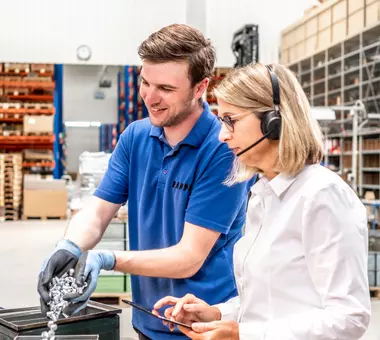 Zwei RAMPA-Mitarbeitende stehen in der Lager-Halle und führen ein fachliches Gespräch. Der Lagermitarbeiter schüttet Muffen in eine Kiste. Die Mitarbeiterin aus dem Kundenservice steht mit Headset auf dem Kopf und Tablet in der Hand daneben.