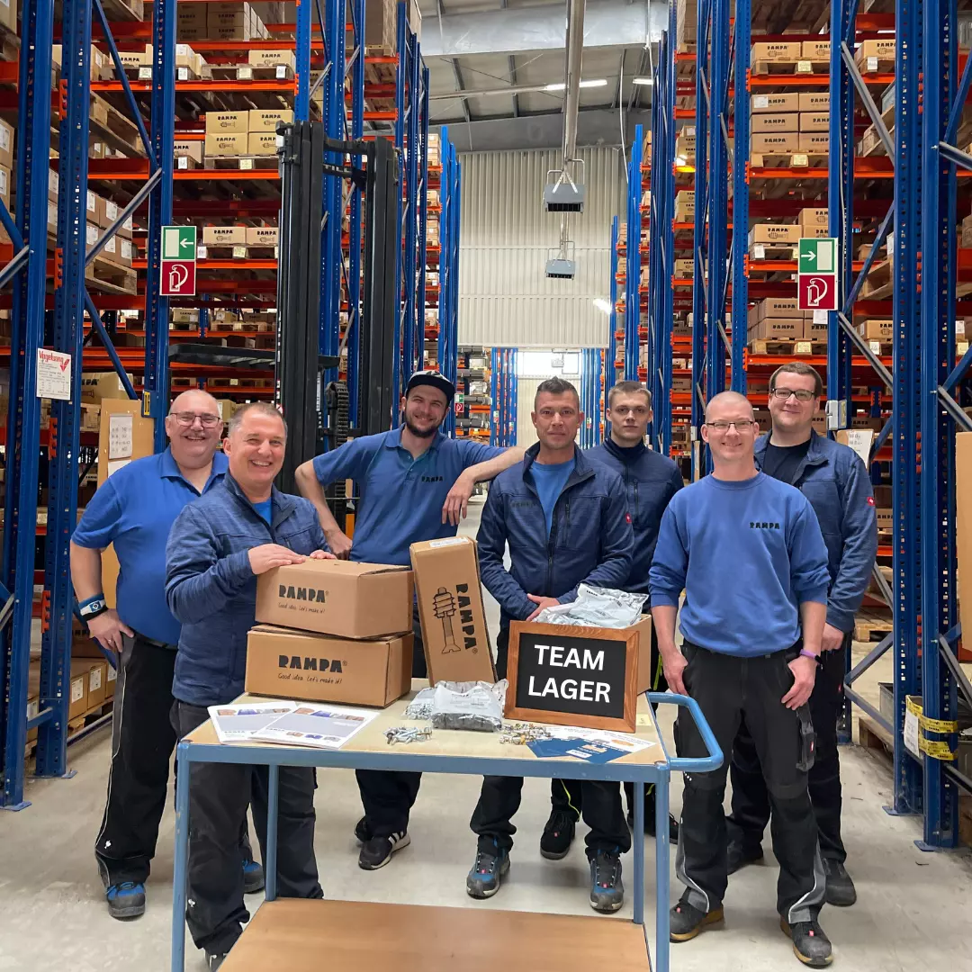 RAMPA team warehouse The warehouse team can be seen in a group picture. In the background are RAMPA's warehouse racks and in the foreground RAMPA cartons, among others, are placed on a packing trolley.