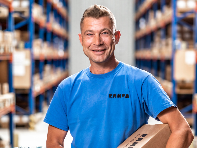 A RAMPA employee representing the warehouse team with a cardboard box in his hand can be seen, with the warehouse in the background.