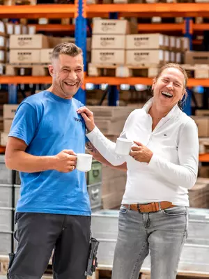 RAMPA's values: We are empathic! Two RAMPA employees are standing next to each other in the warehouse, laughing. Both are holding a cup in their hands. The picture is supposed to stand for RAMPA's value "Empathic".