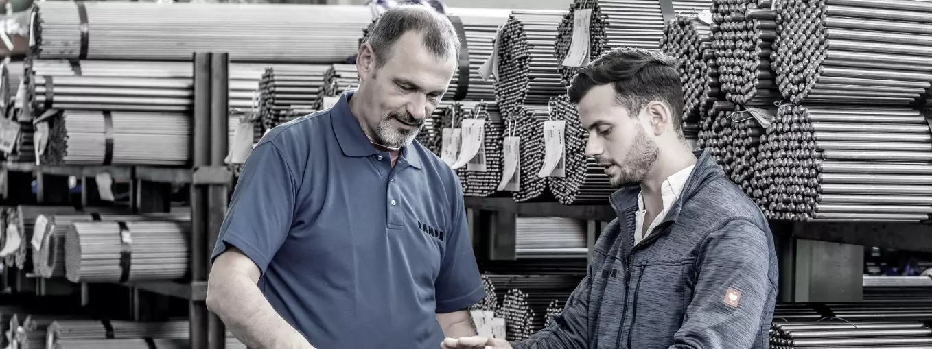 Two RAMPA employees standing in a steel warehouse and exchanging ideas. The picture is meant to symbolize that at RAMPA there is an exchange at eye level across several generations.