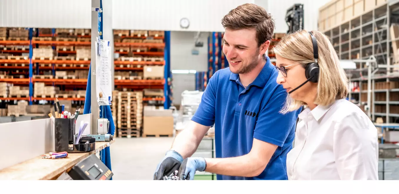 Zwei RAMPA-Mitarbeitende stehen in der Lager-Halle und führen ein fachliches Gespräch. Der Lagermitarbeiter schüttet Muffen in eine Kiste. Die Mitarbeiterin aus dem Kundenservice steht mit Headset auf dem Kopf und Tablet in der Hand daneben.