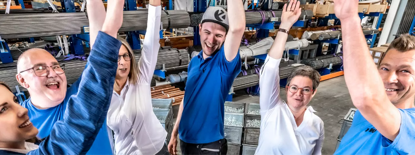 A group of RAMPA employees in a warehouse, standing in a circle and throwing their arms in the air together. The picture is meant to symbolize the high team spirit at RAMPA.
