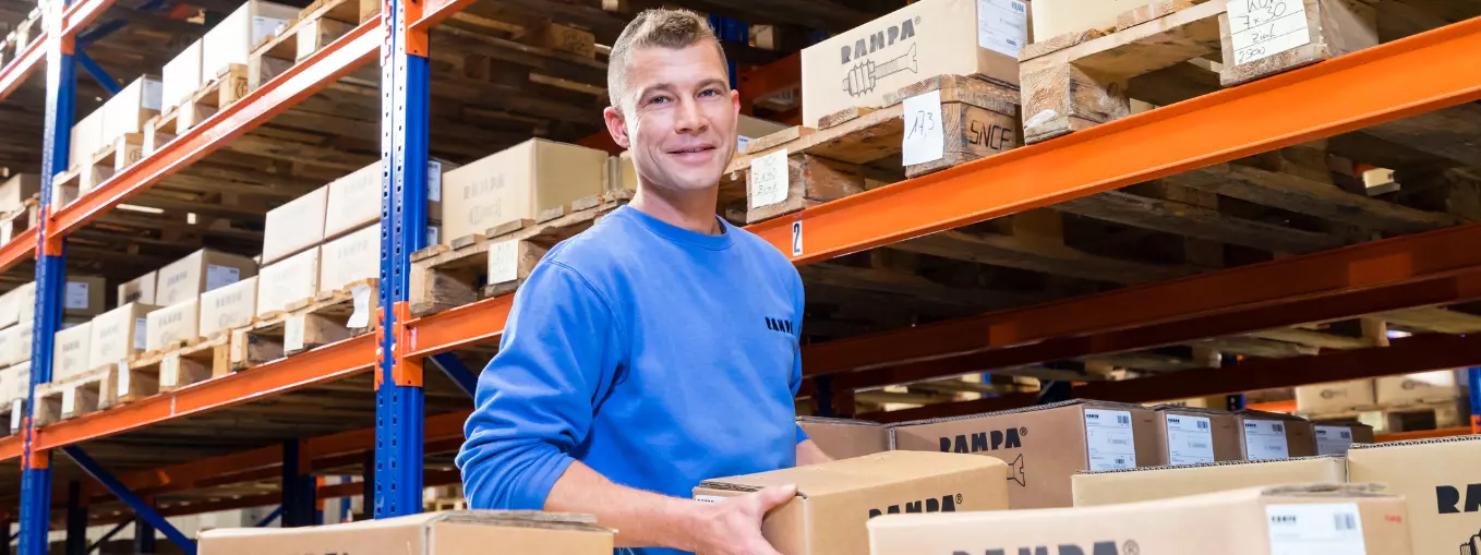 A RAMPA employee from the warehouse holds a RAMPA carton. In front of him are more cartons, behind him is a shelf from the RAMPA warehouse. The picture is meant to reflect our professional performance in the field of service and logistics.